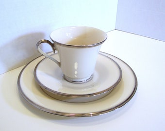 May include: A white teacup, saucer, and bread plate set with silver trim. The teacup has a curved handle and sits atop a matching saucer. A larger bread plate is in the foreground. The set is on a white surface.