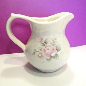 Puede incluir: Jarra de cerámica color crema con asa curva y boquilla. La jarra está decorada con un diseño floral con rosas rosas, pequeñas flores amarillas y hojas verdes. La jarra tiene un cuerpo redondeado y un borde ligeramente ensanchado.