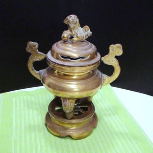 May include: A vintage brass incense burner with a decorative lid featuring a small lion figure. The burner has ornate handles and sits on a round base. The background is a solid black.