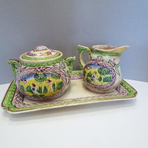 May include: A ceramic tea set with a teapot, creamer, and tray. The set features a colorful pastoral scene with figures and foliage, framed by purple and green accents. The tray is rectangular with a green and purple border.