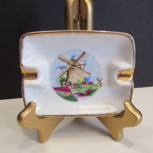 May include: A rectangular white ceramic ashtray with a gold trim and a painted windmill scene. The ashtray has two slots for resting cigarettes. The windmill scene features a brown windmill, green grass, and a blue sky.
