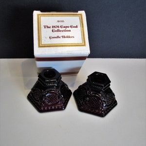 Puede incluir: Dos candelabros de vidrio rojo oscuro con un diseño decorativo. Los candelabros están en su caja original, que dice "Avon The 1876 Cape Cod Collection Candle Holders".