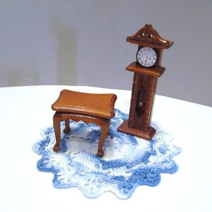 May include: A miniature wooden grandfather clock with a white face and black Roman numerals, paired with a small wooden stool featuring curved legs. Both items are placed on a round, white and blue crocheted doily with a scalloped edge.