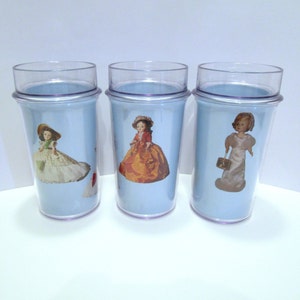 Op de afbeelding: Drie doorzichtige plastic bekers met lichtblauwe zijkanten. Elke beker heeft een vintage poppenafbeelding. De poppen dragen jurken en hoeden. De bekers hebben een doorzichtige plastic boven- en onderkant.