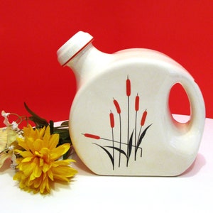 May include: Cream ceramic pitcher with a red rim and handle. The pitcher has a design of red bulrushes with black stems and leaves. A yellow flower and greenery are in the foreground.