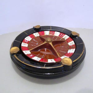 May include: A ceramic roulette wheel with a black exterior, gold accents, and a red, white, and brown interior. The wheel features numbers and is designed for casino games. The wheel has four gold-colored corner pieces.