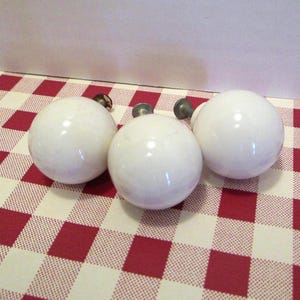 May include: Three white, round cabinet knobs with metal hardware. The knobs are glossy and appear to be ceramic. They are arranged on a red and white checkered tablecloth.