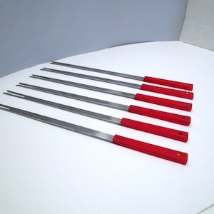 May include: Set of six stainless steel skewers with bright red handles. Each skewer has a two-pronged fork at the tip. The skewers are arranged diagonally across a white surface, suggesting they are ready for use.