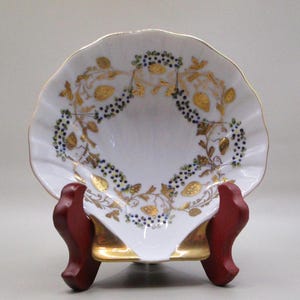 Puede incluir: Un plato de porcelana blanca en forma de concha con un borde dorado y un diseño decorativo de hojas doradas y bayas azules. El plato está sobre un soporte de madera.