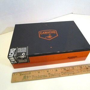 May include: A rectangular cigar box with a black lid and orange base. The lid features the brand name "CAMACHO" in orange, along with a scorpion logo. The box has a sticker that reads "IMPORTED TOBACCO HAND BUILT IN HONDURAS".