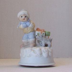 May include: A ceramic figurine of a child in winter attire, carrying gifts with a small dog. The child wears a blue hat, scarf, and pants, a yellow jacket, and boots. The gifts are wrapped with bows.