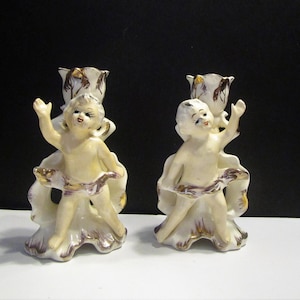 May include: A pair of vintage ceramic candle holders. Each features a cherub figure with an outstretched arm, holding a floral candle cup. The cherubs are cream-coloured with gold and brown accents. The base is white with brown and gold detailing.