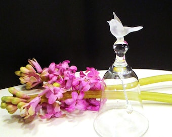 Puede incluir: Una campana de cristal transparente con un pájaro de cristal esmerilado en el mango. La campana tiene un badajo de cadena metálica fina que cuelga en el interior. Junto a la campana hay flores de jacinto rosa intenso con tallos verdes.