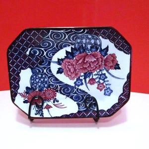 以下が含まれることがあります： 花柄の八角形セラミックプレート。プレートは、青と赤の花柄の白い背景が特徴です。ボーダーは幾何学模様の濃い青色です。プレートは黒い金属製のスタンドに展示されています。