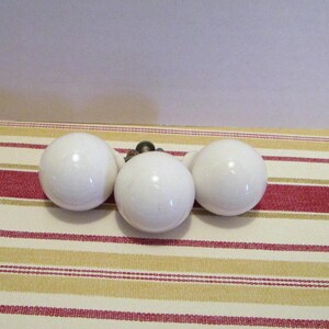 May include: Three white, spherical cabinet knobs with a glossy finish are arranged on a striped fabric. The knobs are evenly spaced, suggesting a set for furniture. The fabric has red, gold, and cream stripes.