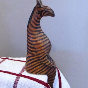 Könnte beinhalten: Eine Holz-Zebra-Figur mit schwarzen und braunen Streifen. Das Zebra sitzt auf einem weißen Handtuch mit rotem Karomuster. Die Figur hat einen langen Hals und ist stilisiert geschnitzt.