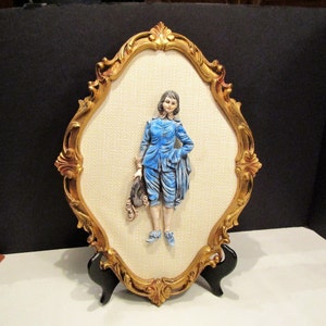 May include: An oval wall hanging featuring a figure in a blue suit, set against a textured beige background. The figure is framed by an ornate gold-colored border. The piece is supported by a black stand.