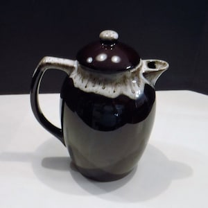May include: A ceramic coffee pot with a dark brown glaze and a white and brown drip pattern around the rim. The pot has a matching lid with a small knob and a curved handle. The spout is on the right side.