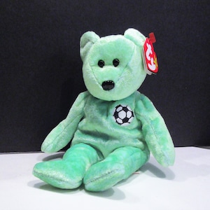 Puede incluir: Un oso de peluche verde claro con un escudo de balón de fútbol en el pecho. El oso tiene ojos negros y una nariz negra. Una etiqueta roja está adherida a su oreja. El oso está sentado sobre una superficie blanca.