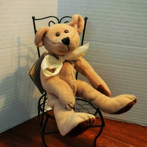 May include: A light tan plush teddy bear with dark brown paw pads and a black stitched nose, wearing a cream-colored satin ribbon tied around its neck. The bear is seated on a small, black, decorative wire chair with a curved backrest.