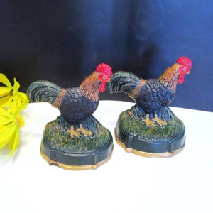 May include: Two decorative rooster figurines. Each rooster has a red comb, orange and brown feathers, and a black body. They are perched on a green and gold base. Yellow flowers are in the background.