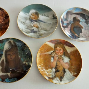 May include: A collection of six decorative plates, each with a gold rim and a unique illustration of children. The plates depict scenes such as a girl with a cat, a child in the snow, and a girl with a bird. The plates are round.