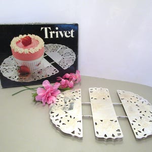 Può includere: Un sottopentola in metallo argentato con un motivo floreale decorativo, accanto a una scatola con la scritta "Trivet" e l'immagine di un dolce. Il sottopentola è in tre pezzi e ha una lunghezza totale di circa 25 cm.