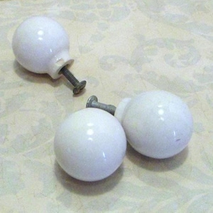 May include: Three spherical white ceramic knobs, each featuring a smooth, glossy finish and a central metal screw attachment extending from the base. These round furniture pulls are designed for installation on cabinets, drawers, or doors.