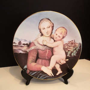 May include: Decorative plate featuring a Renaissance-style painting of a woman and child. The plate has a gold rim and is displayed on a black stand. The artwork depicts a woman in a red dress holding a nude child, with a landscape background.