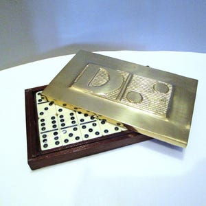 May include: A vintage domino set in a wooden box with a brass lid. The lid has the letter "D" and two circles. The dominoes are white with black dots. The box is open, displaying the dominoes.