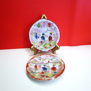 以下が含まれることがあります： 日本のデザインの装飾用セラミックプレート2枚。プレートには、人物、花、提灯のあるカラフルなシーンが描かれています。プレートは、赤い背景に対して金色のスタンドに表示されています。