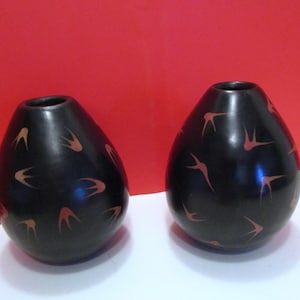 Op de afbeelding: Twee zwarte keramische vazen met een druppelvorm. Elke vaas heeft een smalle opening aan de bovenkant en is versierd met een herhalend patroon van oranje-rode gestileerde vogelvormen. De achtergrond is rood en wit.