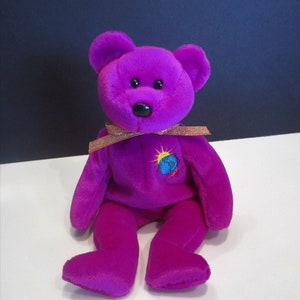 Puede incluir: Un oso de peluche morado con nariz y ojos negros. El oso tiene una cinta dorada alrededor del cuello y un parche bordado de la Tierra con un sol en el pecho. El oso está sentado sobre una superficie blanca.