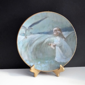 Pode incluir: Prato decorativo com borda dourada, apresentando uma menina de vestido branco e uma gaivota. O prato é exibido em um suporte dourado. A obra de arte usa uma paleta de cores suaves e pastel.