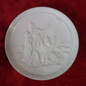 May include: White ceramic plate with a raised relief design depicting the three wise men following a star. The plate has the words "TO THE WISE MEN REJOICE - 02" around the edge. The plate is on a red surface.