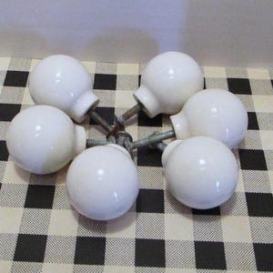 May include: A set of five white spherical cabinet knobs, each featuring a smooth, glossy finish and a central metal screw post extending from the base. The knobs are arranged in a circular pattern with the metal hardware meeting in the center.