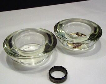 Op de afbeelding: Twee heldere glazen votiefkaarsen, een met een ring erin. Een zwarte ring met een zilveren design ligt ervoor. De kaarshouders staan op een wit oppervlak.