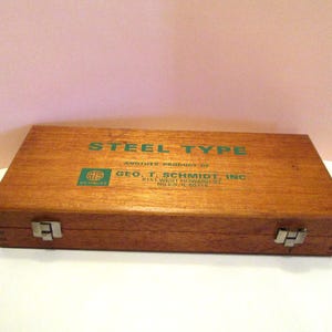 May include: A rectangular wooden storage box with two metal latches on the front. The lid features green printed text that reads STEEL TYPE, ANOTHER PRODUCT OF GEO. T. SCHMIDT, INC. 8151 WEST HOWARD ST. NILES, ILL. 60648 and a green logo.