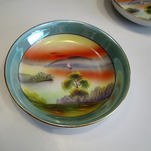 Pode incluir: Duas pequenas tigelas redondas de porcelana com uma cena de paisagem pintada à mão. As tigelas têm uma borda verde-claro com acabamento dourado. O interior apresenta um pôr do sol sobre um lago com um veleiro, árvores e ilhas.