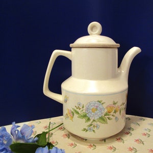 May include: Cream-colored ceramic coffee pot with a lid and handle. The pot features a floral pattern in pastel colors, including blue, yellow, and pink. A blue floral accent is in the foreground. The pot is on a floral patterned surface.