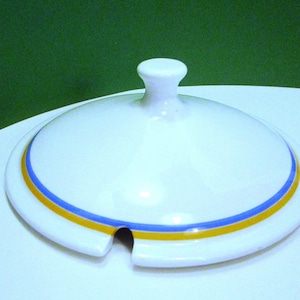 Puede incluir: Tapa de cerámica blanca con forma redondeada y un pequeño asa. La tapa presenta un diseño de rayas azules y amarillas alrededor del borde. Una pequeña muesca está recortada en el lateral. La tapa es probablemente para una fuente o cuenco.