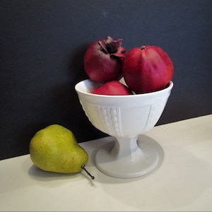 Puede incluir: Naturaleza muerta con un cuenco blanco con pedestal lleno de tres frutos rojos y una pera verde. El cuenco tiene un patrón decorativo. La pera está sobre una superficie blanca, con un fondo negro.