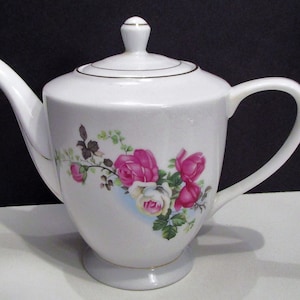 May include: White porcelain teapot with a lid, handle, and spout. The teapot features a floral design with pink and white roses, green leaves, and a gold trim. The teapot sits on a small, round base.