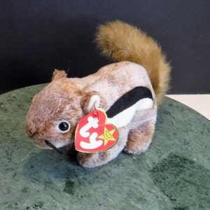May include: A plush squirrel toy with brown fur, a fluffy tail, and a black and white stripe on its side. It has a red heart-shaped tag that reads "Beanie Baby". The toy is sitting on a green surface.