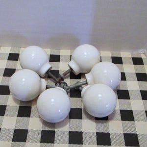 May include: Six white ceramic cabinet knobs with metal hardware are arranged on a black and white checkered surface. The knobs are round with a glossy finish, suggesting a classic or vintage style for home decor.