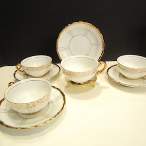 May include: A white porcelain tea set with gold trim. The set includes four teacups with matching saucers and a serving plate. The cups have gold handles and decorative gold patterns. The saucers and plate have gold rims and gold line accents.