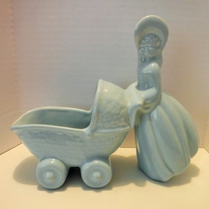May include: A light blue ceramic figurine of a woman pushing a baby carriage. The woman is wearing a bonnet and a long dress. The carriage has four wheels and a curved hood. The figurine is a vintage style.