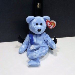 May include: A light blue plush bear with a snowflake pattern and a dark blue scarf. The bear has black eyes and a black nose. A red and white tag is attached to the bear's ear.