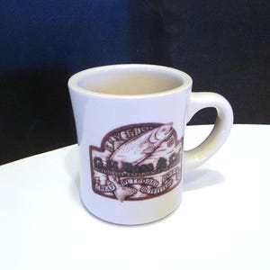 May include: Cream-colored ceramic mug with a curved handle. The mug features a brown oval design with a fish illustration and the text "Fly Fishers Great Outdoor Outfitters". The mug is designed for beverages.