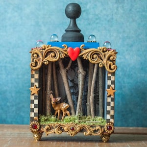 Könnte beinhalten: Eine dekorative Box mit einer Waldszene. Die Box zeigt eine Hirschfigur, Miniaturbäume und ein rotes Herz. Der Rahmen ist mit goldenen Akzenten, einem schwarzen Schachmuster und einem schwarzen Bauern oben verziert. Der Hintergrund ist eine blaue Wand.